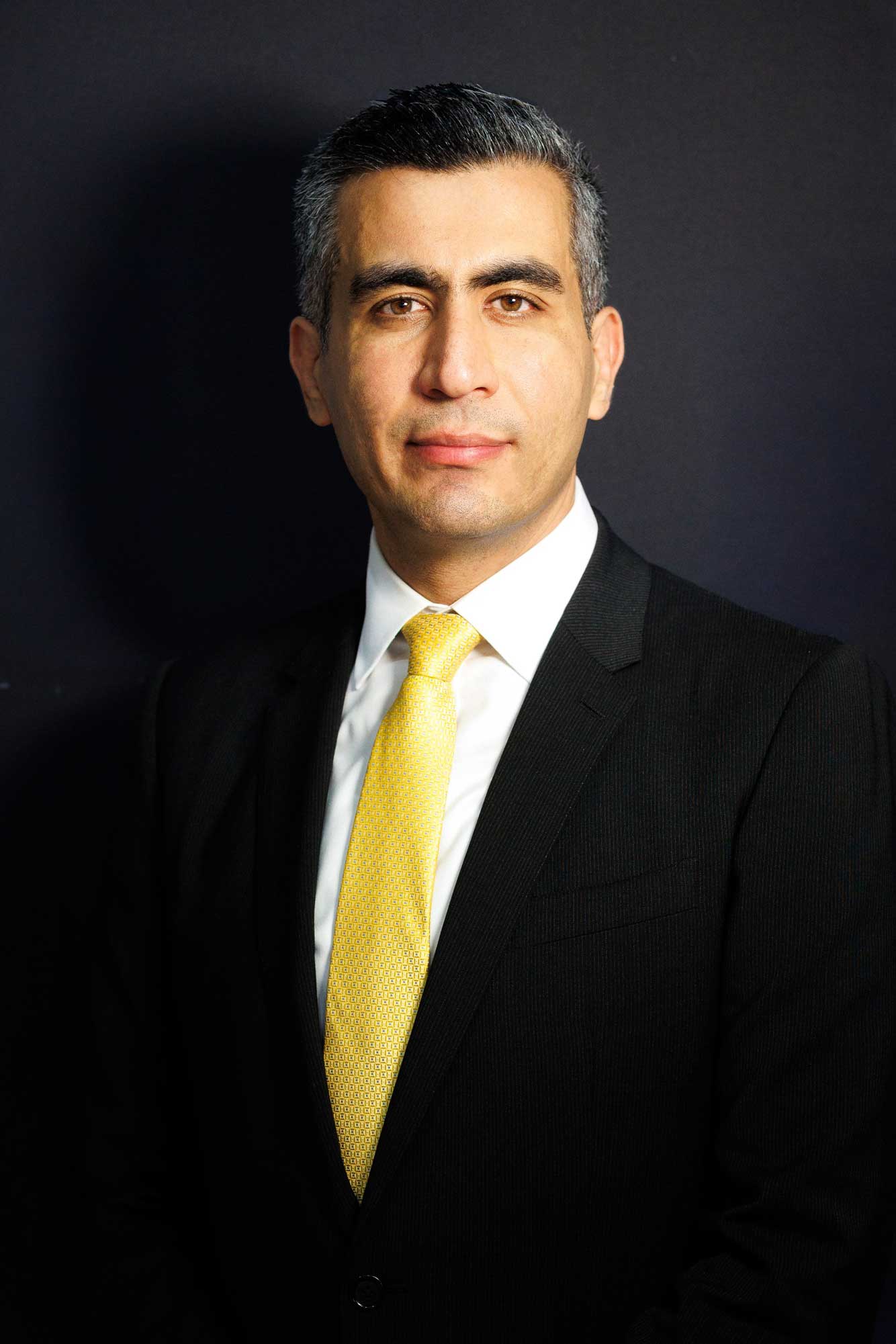 Ali Aydan in a formal black suit, white shirt, and yellow tie, posing for a professional studio portrait against a dark background.