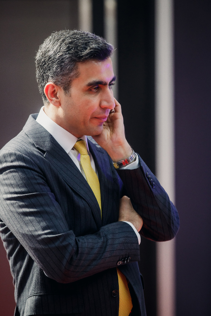 Ali Aydan in a pinstripe suit with a yellow tie speaking at a business event.